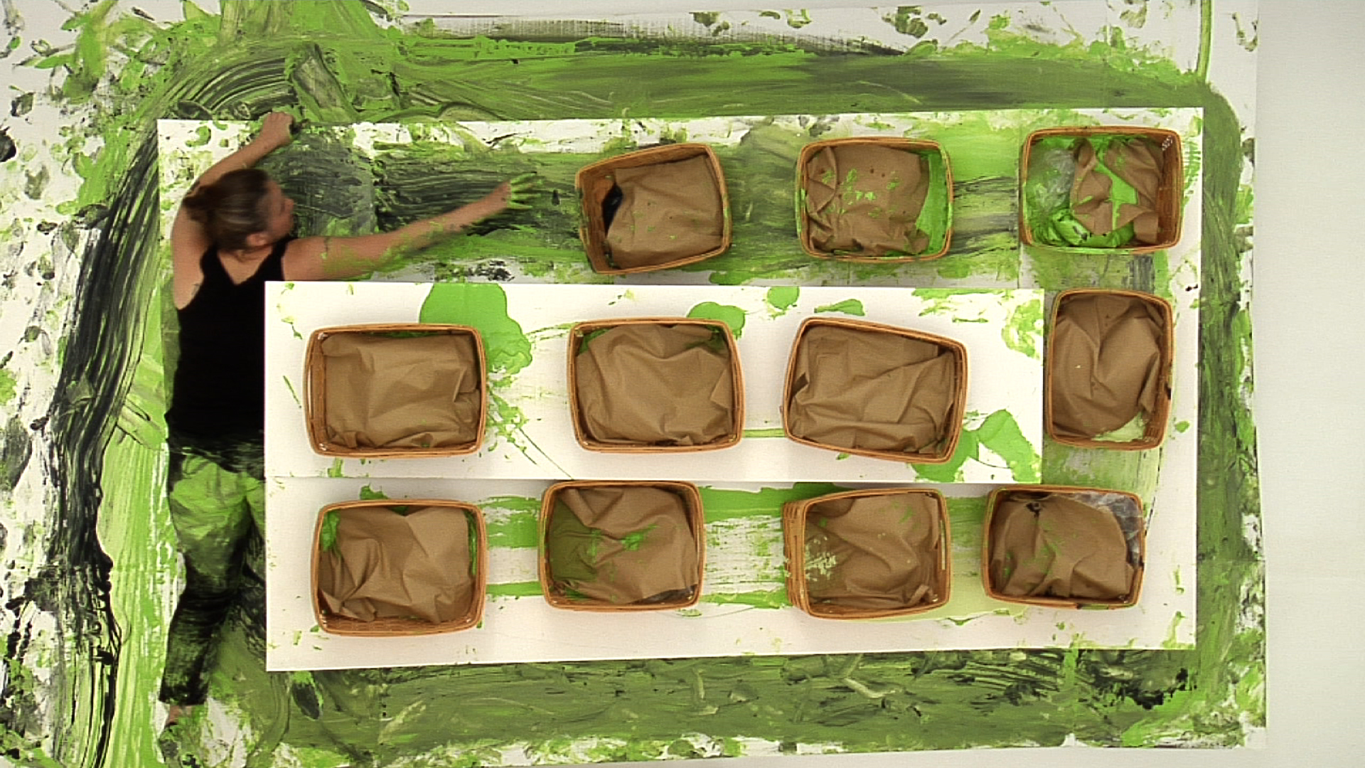 Aerial view of 11 baskets and a woman wearing all black on a white platform. Green and black paint is smeared on the platform and is also on the woman and baskets.