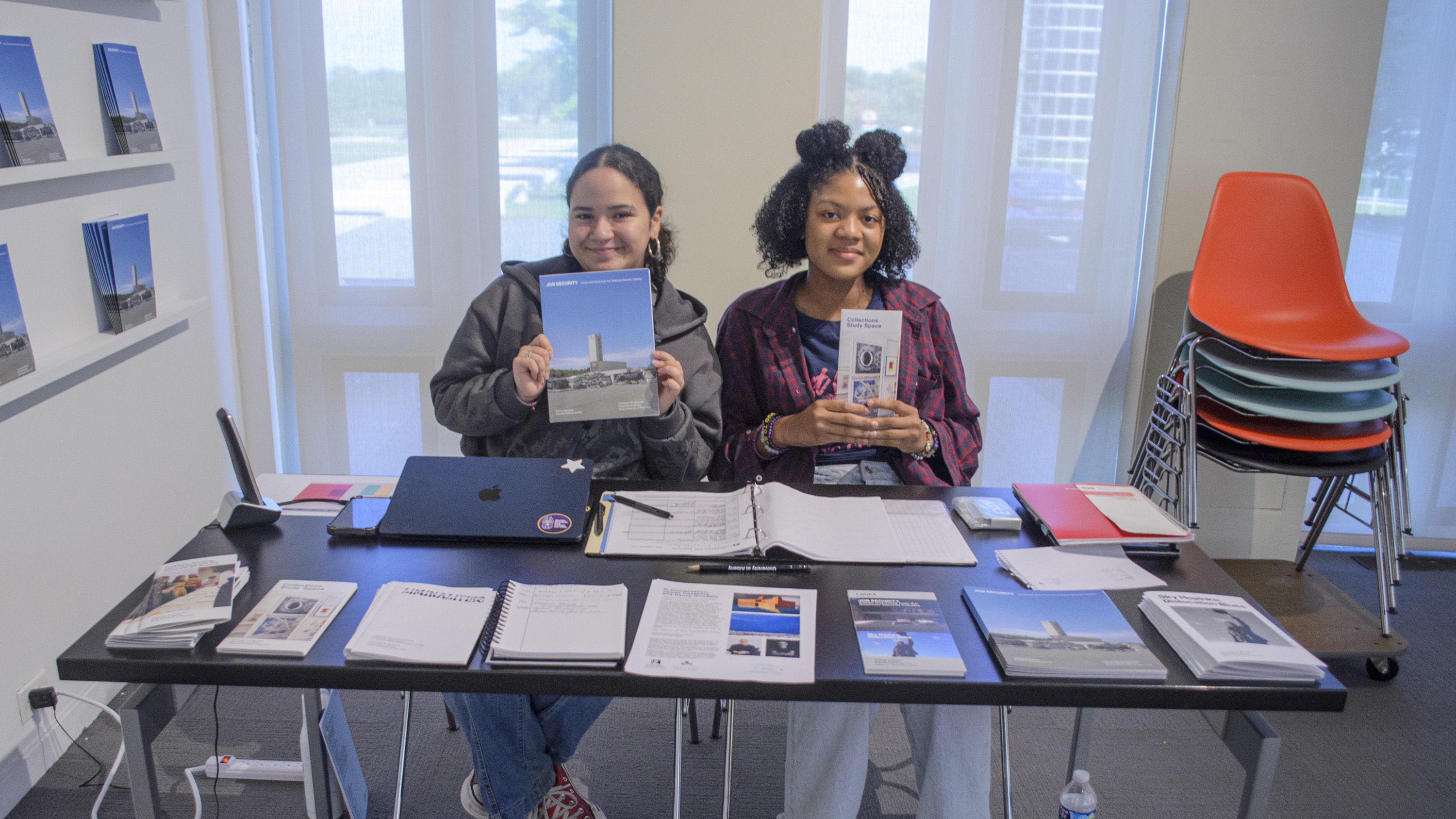 Two students sitting at a desk holding up brochures and smiling. The desk has additional brochures on it.