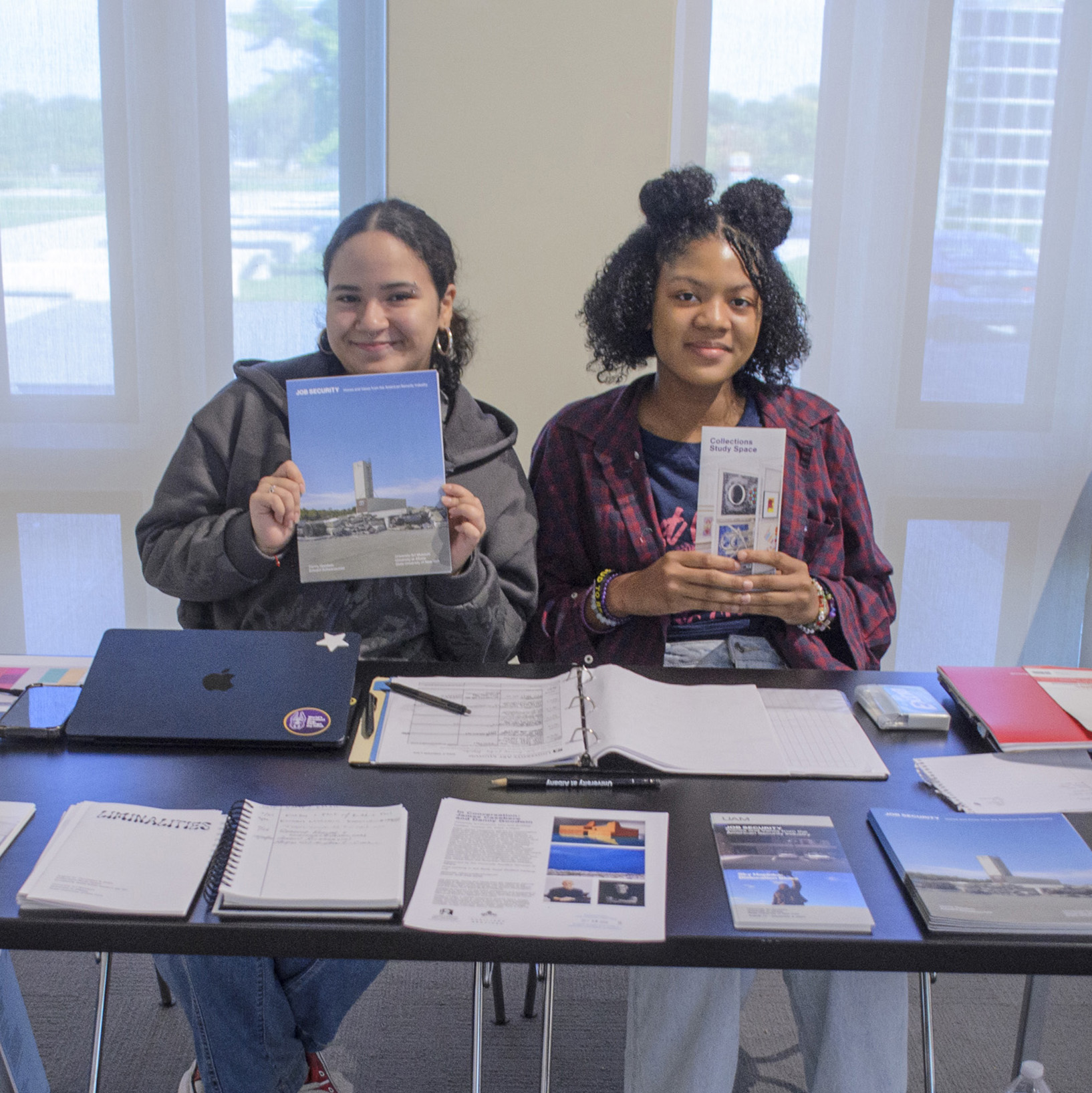 Two students sitting at a desk holding up brochures and smiling. The desk has additional brochures on it.