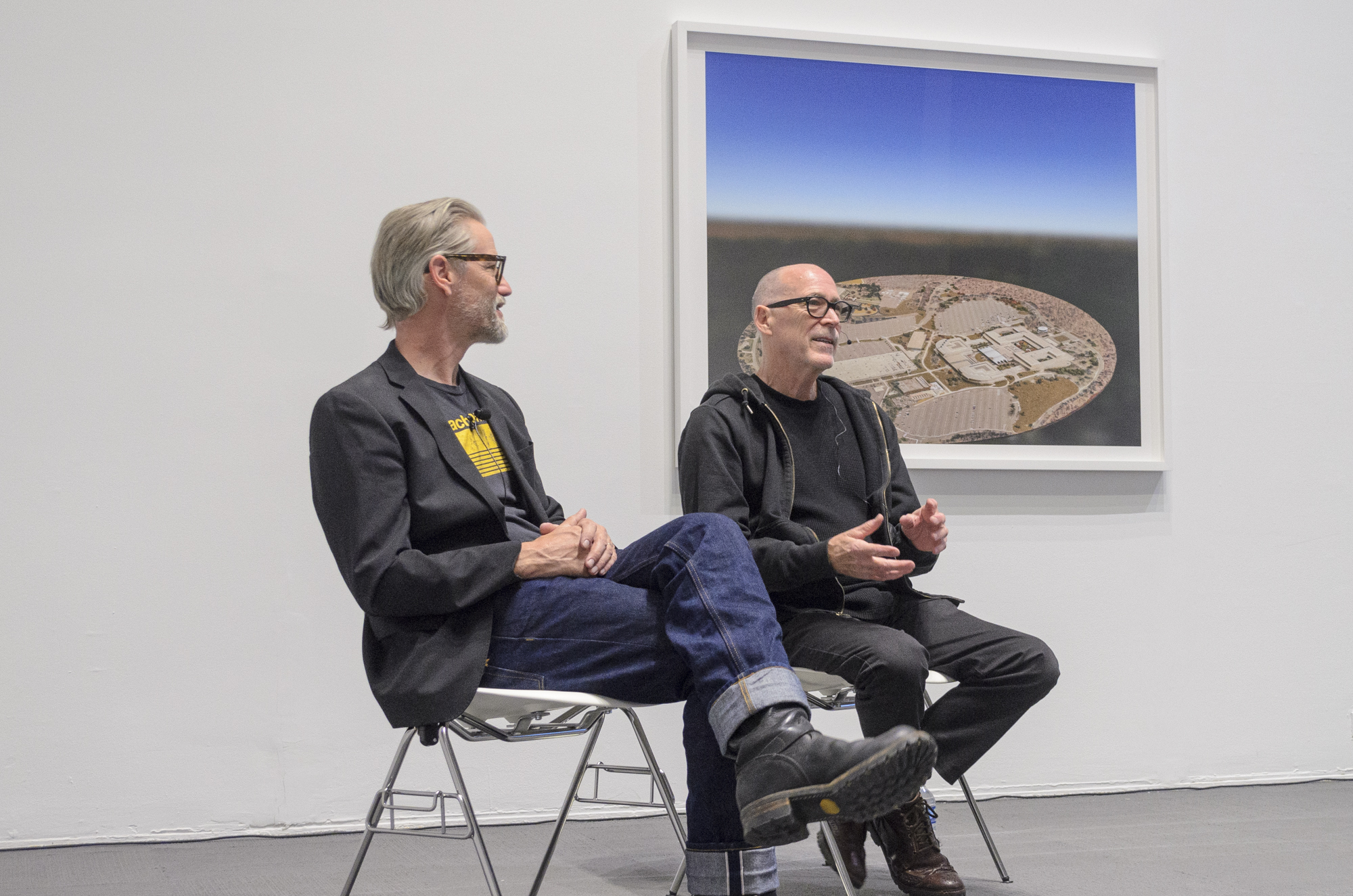 Two men seated and engaged in conversation in front of a framed photograph.