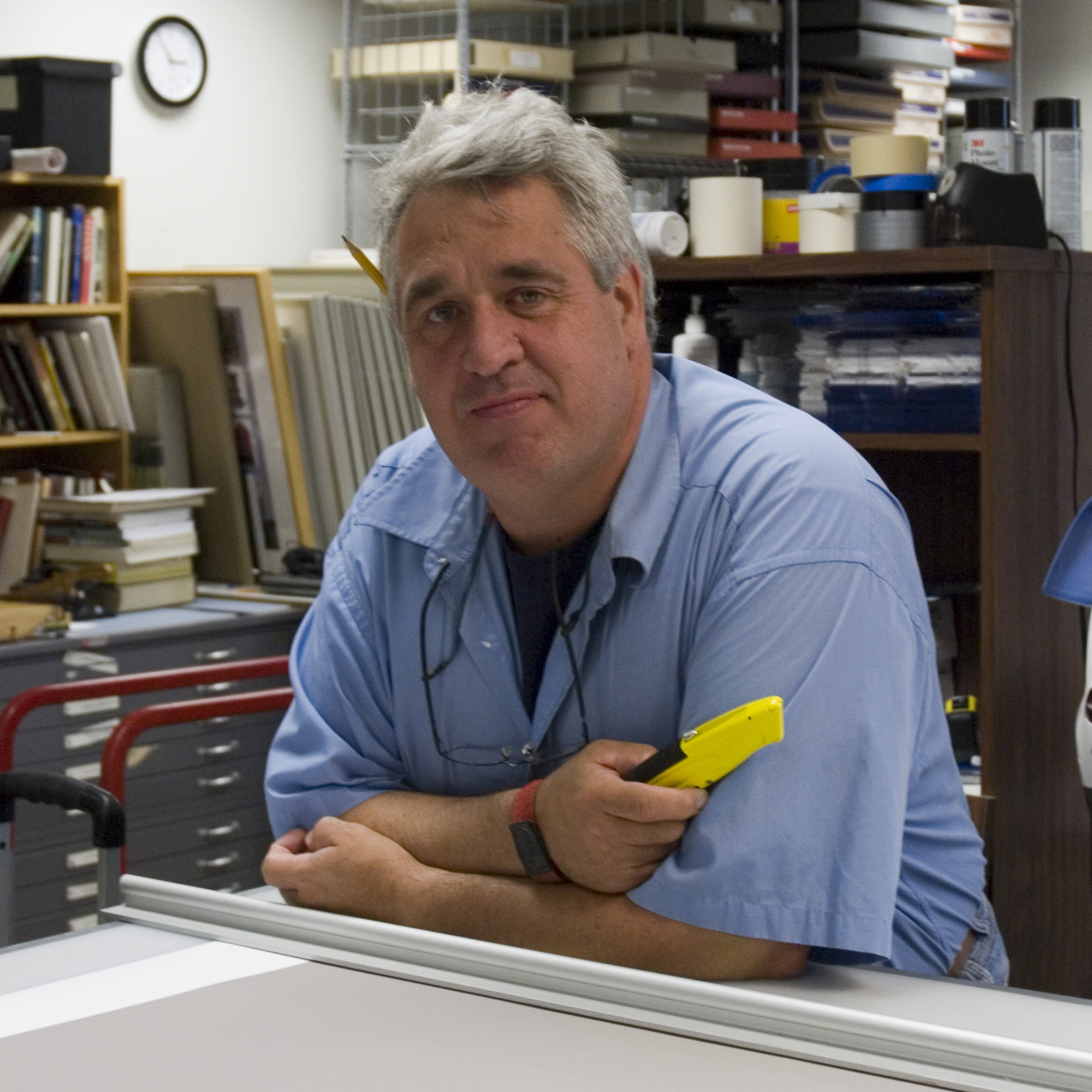 A man in a blue shirt leaning on a table, holding a yellow utility knife.