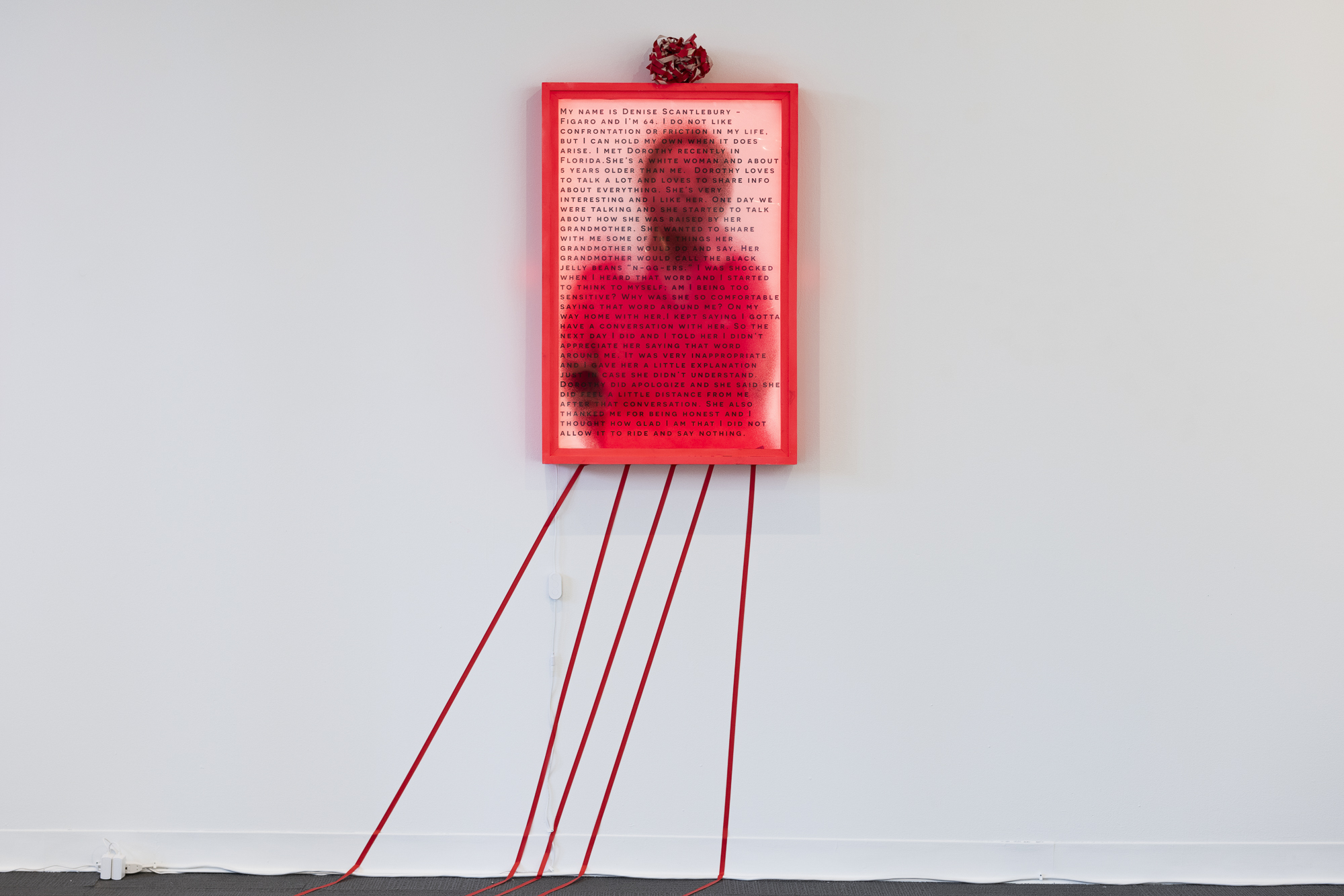 A blurry image of a person wearing red with text overlaid on  the image is framed in a bright red frame. A ball of red tape is on top of the frame, and tape lines are traveling down from the frame to the floor.